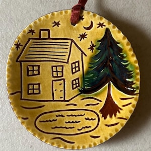 May include: A round, yellow ceramic ornament with a scalloped edge and a dark red ribbon hanger. The surface features a hand-painted scene of a house, a tall evergreen tree, a small pond, and a night sky with stars and a crescent moon.