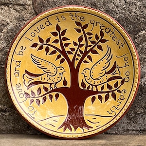 May include: A decorative ceramic plate with a yellow background and a brown tree design. Two birds perch on the tree branches. The plate's rim is brown, and the text "Love and be loved is the greatest joy on earth" is written around the edge.