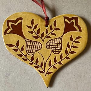 May include: A heart-shaped ceramic ornament with a mustard yellow glaze and dark brown painted details. The design features two stylized flowers with grid-patterned centers and leafy stems. A thin red ribbon is attached to the top for hanging.