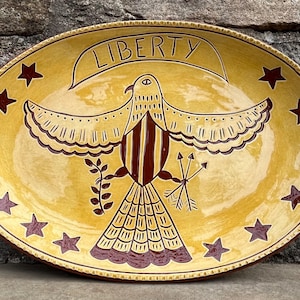May include: Oval, hand-painted ceramic platter with a yellow background and a central design of a bird with outstretched wings. The word "LIBERTY" is above the bird. Dark red stars and accents complete the design.