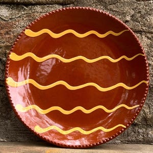 May include: A round, glazed terracotta plate featuring a deep reddish-brown base color. The surface is decorated with five horizontal, wavy yellow lines. The outer edge of the plate has a distinct, textured, scalloped or beaded rim detail.