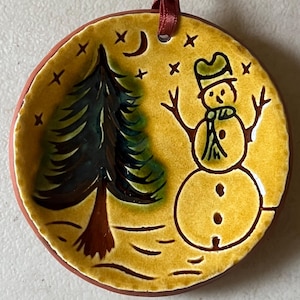 May include: A round ceramic ornament with a yellow glazed surface featuring a hand-painted dark green pine tree and a snowman wearing a green hat and scarf. The design includes a crescent moon and stars, with a red ribbon loop at the top for hanging.