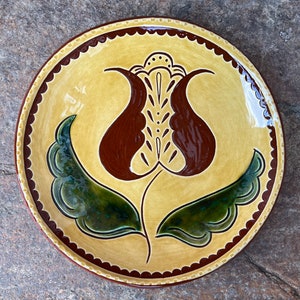 May include: A yellow ceramic plate with a brown and green floral design. The plate has a scalloped edge and a brown rim.