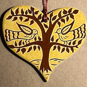May include: A heart-shaped ceramic ornament with a yellow glaze and a dark brown painted design of a central tree with two birds perched on its branches. A thin dark red ribbon is attached to the top for hanging this decorative piece.