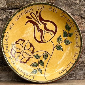 May include: A yellow ceramic plate with a floral design and text. The plate features a large brown tulip and a smaller flower, with green leaves and a brown border. The text is in a script font.