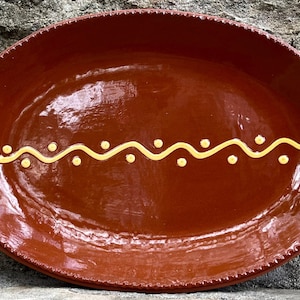 May include: An oval-shaped terracotta serving platter with a glossy brown glaze. The rim features a textured, beaded edge. A horizontal yellow wavy line with small yellow dots positioned above and below the curves decorates the center.