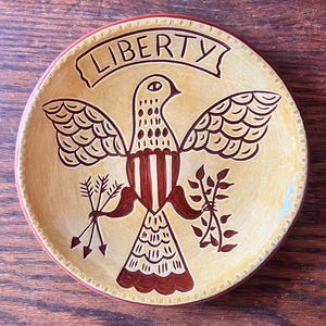 Peut inclure: Une assiette décorative en céramique avec un motif patriotique. L'assiette présente une image centrale d'un oiseau stylisé aux ailes déployées, tenant des flèches et une branche. Le mot "LIBERTY" est affiché en haut. L'assiette a un fond jaune avec des accents marron et rouge.