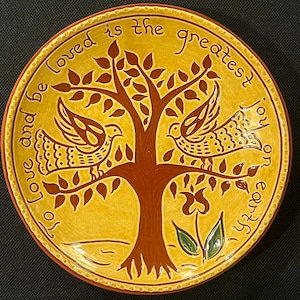 May include: A round, ceramic plate with a yellow background and a brown tree design. Two birds are perched on the tree branches. The text "Love and be loved is the greatest on earth" is written around the edge of the plate.