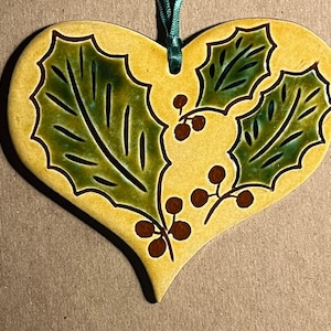 May include: A heart-shaped ceramic ornament with a yellow glazed finish, featuring a hand-painted design of green holly leaves and brown berries. A teal ribbon is attached to the top for hanging, providing a decorative accent to the piece.