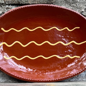May include: An oval-shaped terracotta serving platter with a glossy, deep reddish-brown glaze. The surface features three horizontal, wavy yellow lines painted across the center. The rim is decorated with a raised, beaded texture pattern.