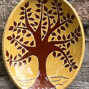 May include: An oval ceramic platter with a glossy mustard yellow glaze featuring a hand-painted dark brown tree design. The tree has a thick trunk and spreading branches with numerous leaves. The rim of the platter has a textured, notched edge.
