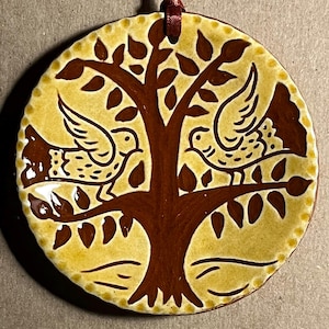 May include: A circular ceramic ornament featuring a glossy yellow background with a dark brown painted design of a central tree and two birds perched on its branches. The edges of the ornament have a scalloped texture and a dark ribbon is attached.