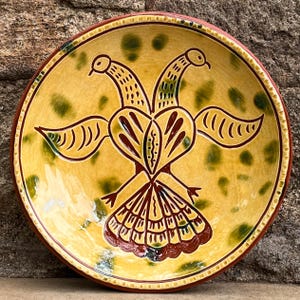 May include: A hand-painted ceramic plate with a yellow background and a central design of a two-headed bird in brown and green. The plate has a red rim and green accents, showcasing a folk art style.
