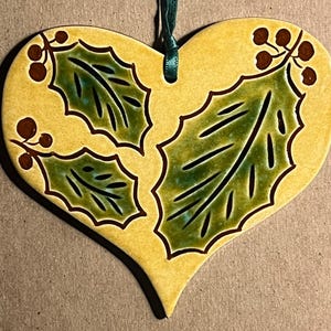 May include: A heart-shaped ceramic ornament with a yellow glazed finish, featuring hand-painted green holly leaves with dark brown outlines and clusters of brown berries. A dark green ribbon is threaded through a hole at the top for hanging.