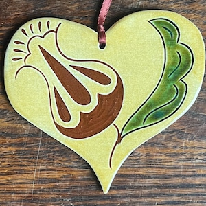 May include: A heart-shaped ceramic ornament with a glossy pale yellow glaze. The surface features a hand-painted brown stylized flower design on the left and a dark green leaf on the right. A thin maroon ribbon is threaded through a hole at the top.
