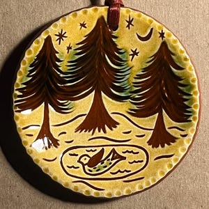 May include: A round ceramic ornament featuring a hand-painted scene of three brown pine trees against a yellow background with stars and a crescent moon. A small bird is depicted in a pond at the bottom. A dark red ribbon is attached to the top.