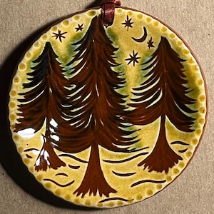 May include: A round ceramic ornament featuring a hand-painted forest scene with three dark brown pine trees against a yellow background. The design includes a crescent moon and stars at the top, a dotted border, and a dark red ribbon for hanging.