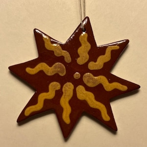 May include: A star-shaped ornament with a dark red background and yellow squiggly designs. The ornament is suspended by a thin, off-white string. The star has eight points and a glossy finish.