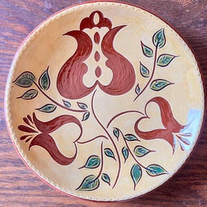 May include: Circular ceramic plate with a beige background and a floral design in brown and green hues. The rim has a dotted pattern and a red border. The floral design features stylized flowers and leaves.