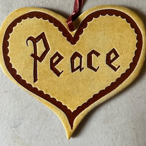 May include: A heart-shaped ceramic ornament in a light tan color with a dark brown scalloped border. The word "Peace" is embossed in the center in a dark brown font. A thin, dark red ribbon is attached to the top for hanging purposes.
