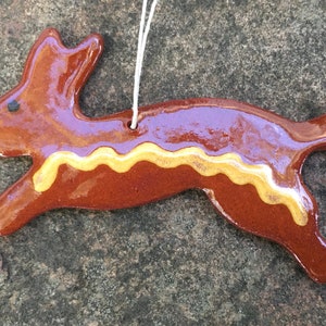 May include: A ceramic ornament in the shape of a leaping rabbit. The ornament is brown with a yellow wavy line along the belly and a white string for hanging. The rabbit has a glossy finish and a small black dot for an eye.