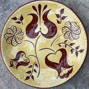 May include: A decorative ceramic plate with a yellow background and a brown floral design. The plate features stylized flowers, leaves, and a brown rim. The design is symmetrical and the plate has a textured edge.