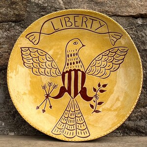 Peut inclure: Une assiette ronde en céramique jaune avec un motif d'oiseau marron. L'oiseau a les ailes déployées et tient des flèches et une branche. Le mot "LIBERTY" est écrit en haut en marron.