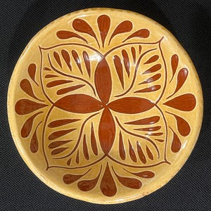 May include: A small, round, ceramic bowl with a light yellow base and a symmetrical, floral-inspired design in shades of brown. The bowl's interior features a central four-petal flower design, surrounded by stylized leaf shapes.
