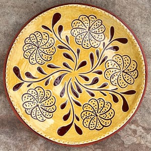 May include: A round, hand-painted ceramic plate with a yellow background and a dark brown floral design. The plate features five stylized flowers and decorative leaf-like shapes. The rim is a darker brown color.