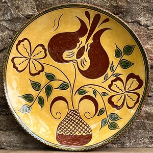 May include: A hand-painted ceramic plate with a yellow background and a floral design. The design features brown flowers and a vase, with green leaves and a green border. The plate is round and has a rustic, artisanal look.