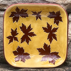May include: Square ceramic serving dish with a light yellow interior and a dark brown rim. The dish is decorated with dark brown leaf patterns. The background is a textured stone surface.