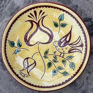 May include: A decorative ceramic plate with a hand-painted design. The plate features a yellow background with a brown rim and a floral and bird motif in brown, green, and purple. The plate's diameter is approximately 10 inches.