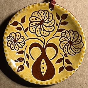 May include: A round ceramic ornament with a glossy mustard yellow glaze and a scalloped edge. The surface features a hand-painted dark brown design of a central vase holding three stylized flowers with dotted petals and leafy stems.