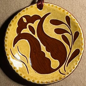 May include: A round ceramic ornament featuring a glossy mustard yellow glaze with a dark brown painted floral motif. The edge has a raised dotted texture, and a dark red ribbon is threaded through a hole at the top for hanging purposes.