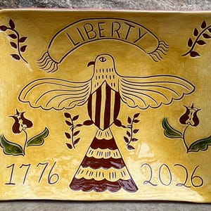 May include: A rectangular, yellow ceramic platter with a hand-painted design. The design features a stylized eagle with outstretched wings, a banner reading "LIBERTY", and the dates "1776" and "2026". Decorative floral and leaf elements complete the design.