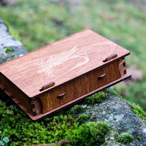 May include: A wooden box with a drawer, featuring a laser-etched design of a fly on the lid. The box is sitting on a mossy rock.