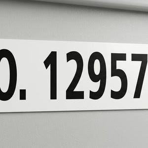 May include: A rectangular white sign with rounded corners displays the text "NO. 1295734" in bold black font. The sign is mounted on a light gray wall.