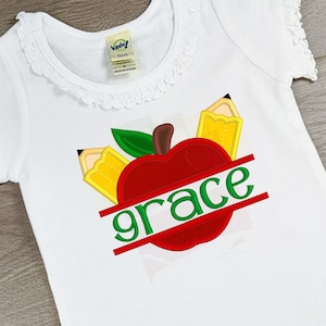 Puede incluir: Camiseta blanca con una manzana roja, hojas verdes y dos lápices amarillos. La palabra "grace" está escrita en letras verdes sobre la manzana.