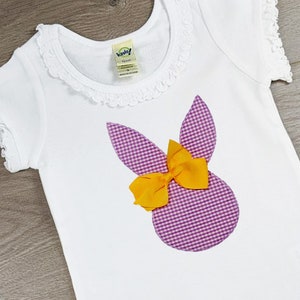 May include: White short-sleeved t-shirt featuring a purple and white gingham bunny design with a yellow bow. The shirt has a frilled collar and sleeve edges. The tag reads "Vando!"