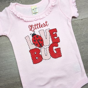 Pode incluir: Um body de bebê rosa claro com as palavras "Littlest Love Bug" bordadas em fio vermelho e rosa. Uma joaninha é bordada no centro da palavra "Love". O body tem uma gola com babados e mangas curtas.