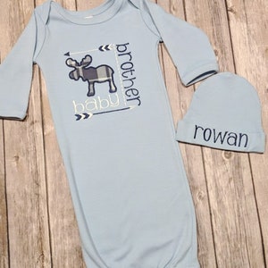 May include: A light blue baby gown and matching beanie set. The gown features an embroidered moose with a plaid pattern and the words "baby brother". The beanie is embroidered with the name "rowan".