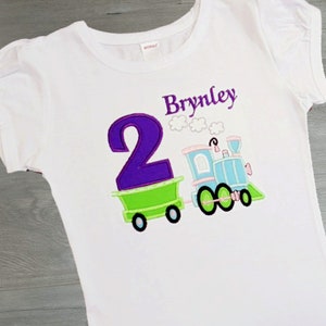 Puede incluir: Camiseta blanca con un diseño de tren colorido y el número 2. El tren es azul claro con ruedas negras y un vagón verde. El nombre "Brynley" está bordado en morado encima del tren.