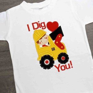 Puede incluir: Camiseta blanca con un gráfico colorido de un vehículo de construcción amarillo con un personaje de dibujos animados en su interior. El texto "I Dig You!" es rojo.