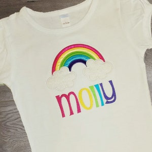 May include: White t-shirt with a rainbow and clouds embroidered on the front. The word "molly" is embroidered below the rainbow in colourful letters.