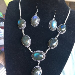 Puede incluir: Un conjunto de collar y pendientes de plata con piedras de labradorita azul verdosa iridiscente de forma ovalada. El collar tiene cinco piedras y un colgante, mientras que los pendientes tienen una piedra cada uno.