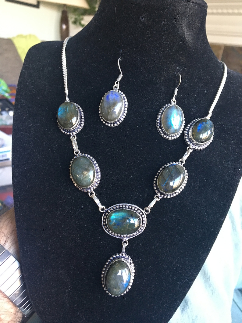 Puede incluir: Un conjunto de collar y pendientes de plata con piedras de labradorita azul verdosa iridiscente de forma ovalada. El collar tiene cinco piedras y un colgante, mientras que los pendientes tienen una piedra cada uno.