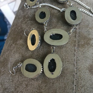 Puede incluir: Un conjunto de collar y pendientes de plata con piedras de forma ovalada. Las piedras son de color verde oscuro y tienen un engaste de plata. El collar tiene un dise&ntilde;o de cadena de eslabones.