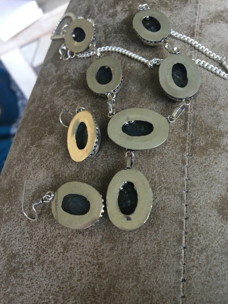 Puede incluir: Un conjunto de collar y pendientes de plata con piedras de forma ovalada. Las piedras son de color verde oscuro y tienen un engaste de plata. El collar tiene un dise&ntilde;o de cadena de eslabones.