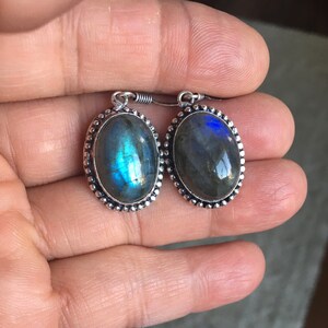 Puede incluir: Un par de pendientes de plata con piedras de labradorita de forma ovalada. Las piedras tienen una base gris oscura con destellos iridiscentes azules y verdes.