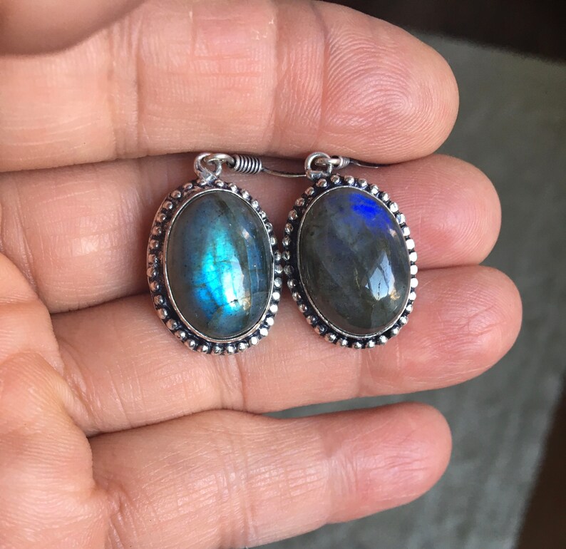 Puede incluir: Un par de pendientes de plata con piedras de labradorita de forma ovalada. Las piedras tienen una base gris oscura con destellos iridiscentes azules y verdes.